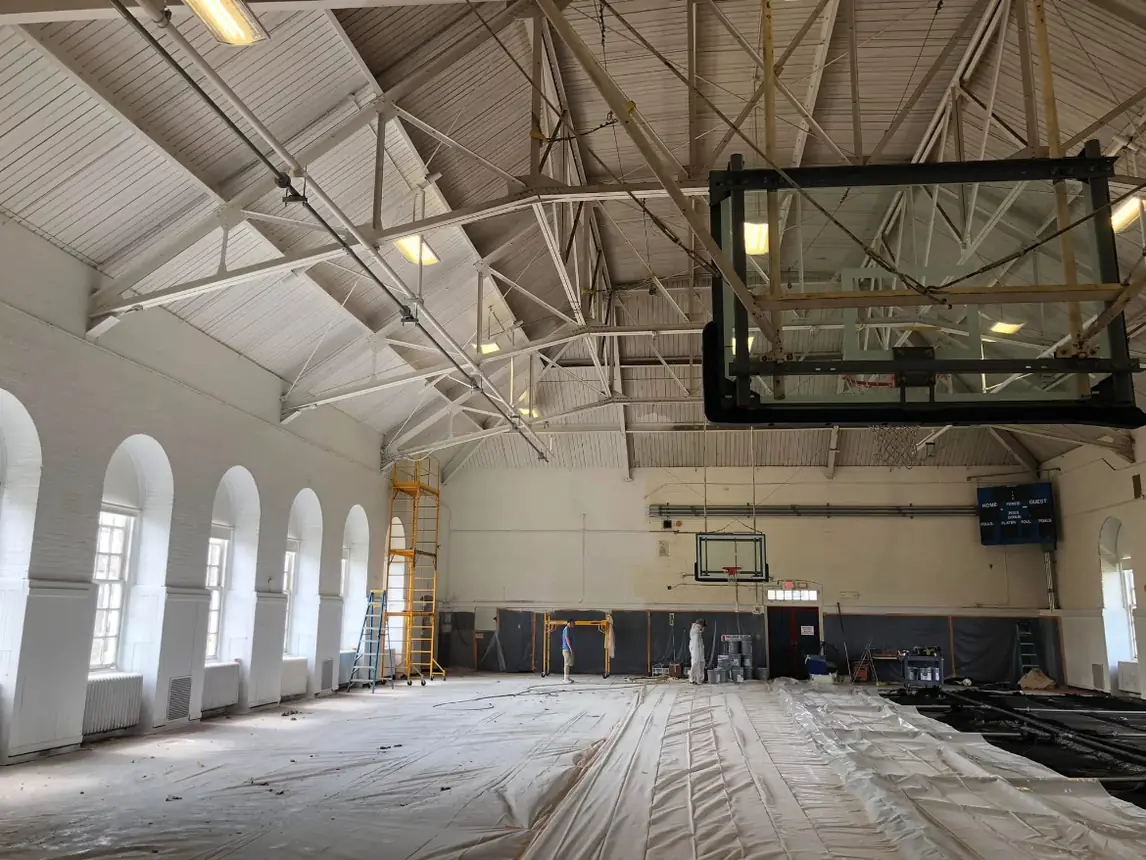 Gymnasium floor fully covered in drop cloths with plastic sheeting over the lower walls partway through the repaint