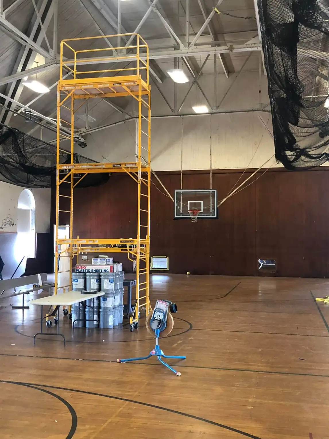 Gymnasium before painting with a rolling scaffold tower set up, a dark stained end wall, and paint supplies staged on the floor