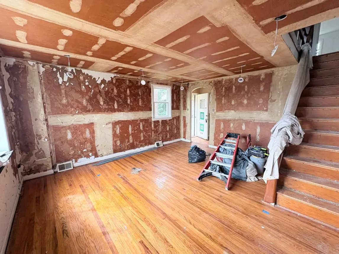 Living room with wallpaper stripped off, bare patched plaster on the walls and ceiling, and a ladder and debris bags on the hardwood floor
